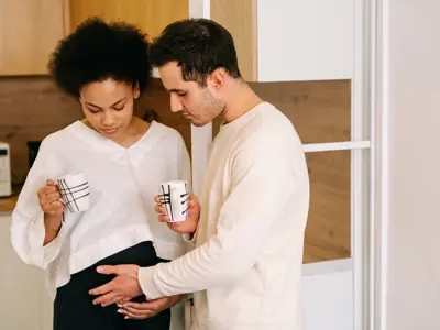 A couple stands together in a kitchen - the woman, who appears to be pregnant, looks down at her belly while the man gently places his hand on it, showing a moment of connection.