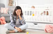 Woman holding her head in her hands, sitting beside a crib with a baby. 