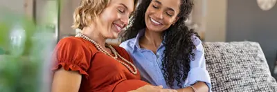 A same sex couple sitting on a couch gazing at the pregnant partner's belly.