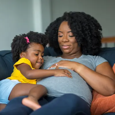 Mom and toddler daughter sitting on a couch with their hands on the Mom's pregnant belly.